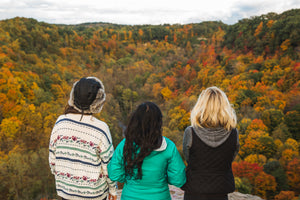 3 women sitting on a cliff overlooking forest of fall colored trees